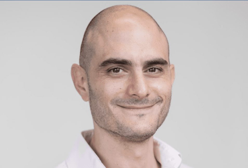 A man with a bald head, wearing a light pink shirt, smiles at the camera against a neutral background.